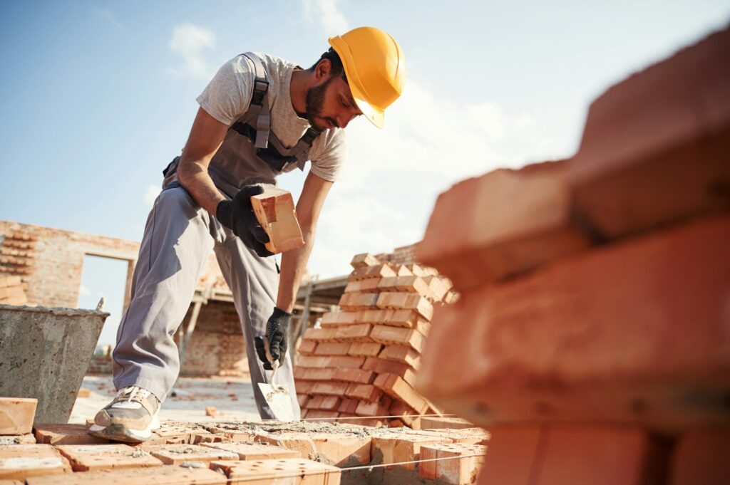 View from below. Brick in hand. Handsome Indian man is on the construction site